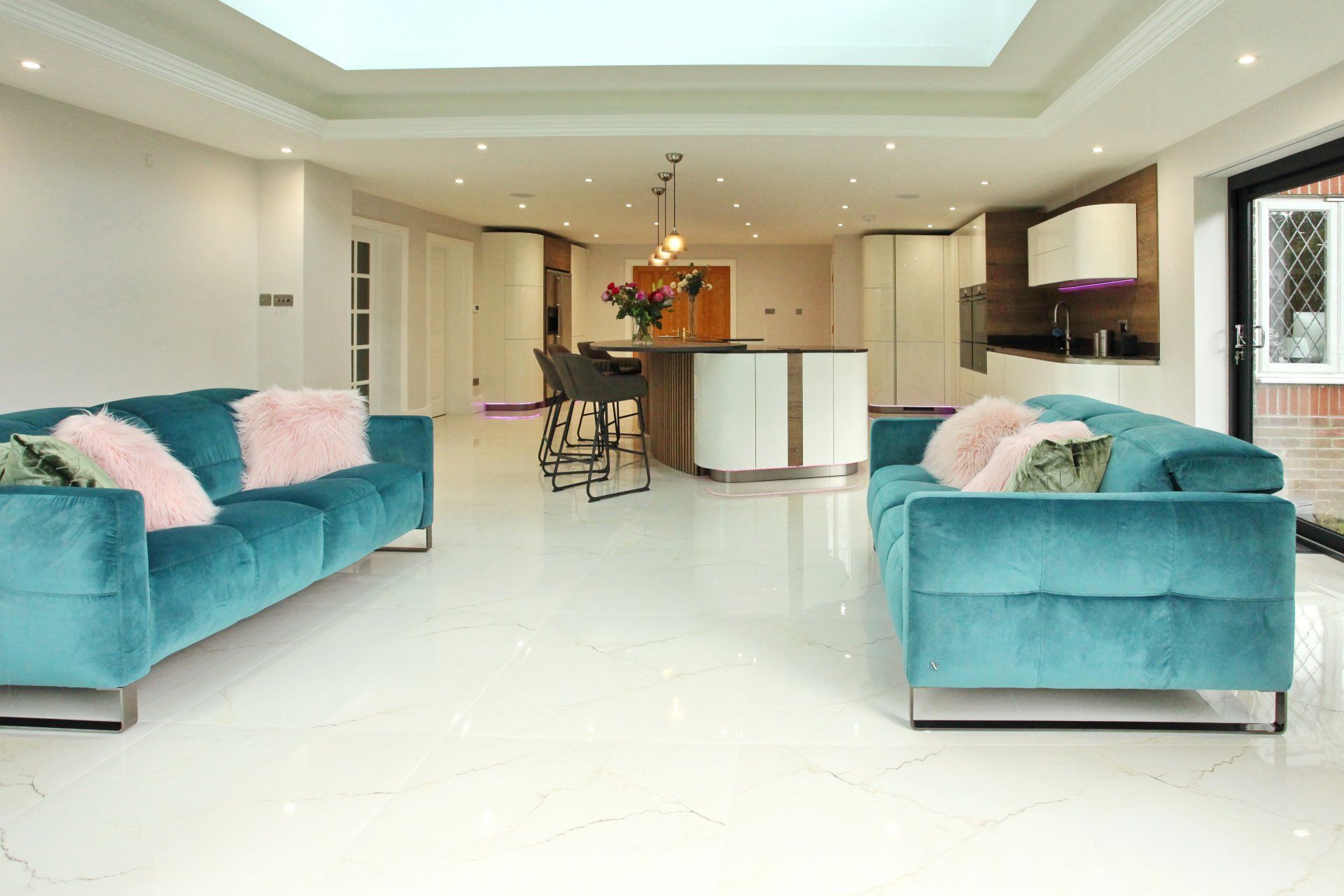 Spacious living room with turquoise velvet sofas, pink pillows, and a modern kitchen. White floors and walls.