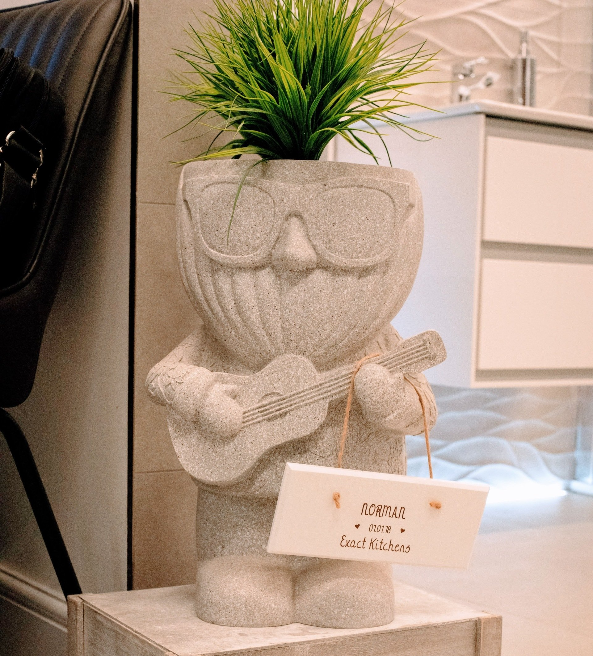 A stone-like planter shaped like a musician with glasses, playing guitar, holding a sign; green plant in its head.