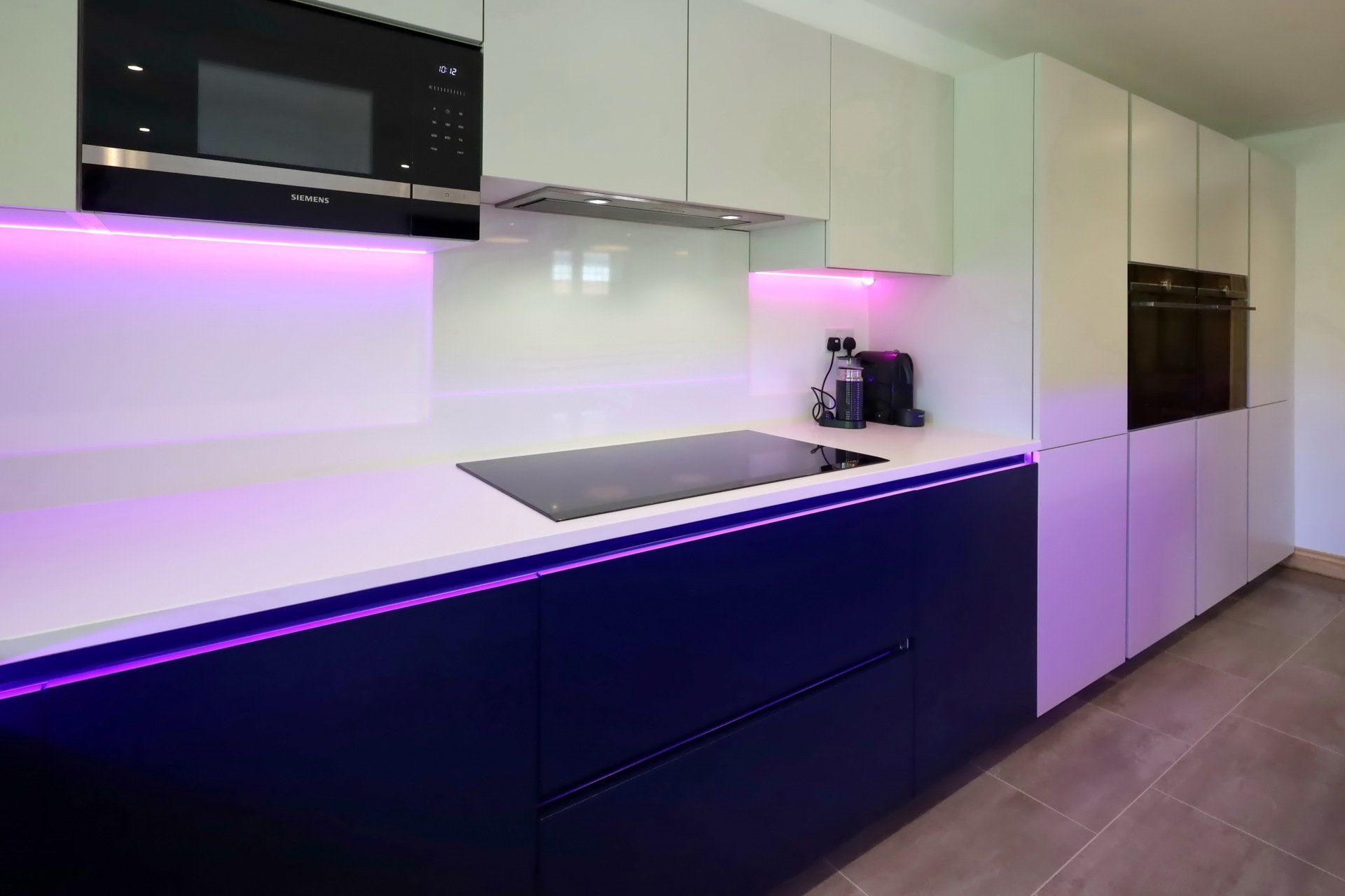 Modern kitchen with white and dark blue cabinets, purple lighting, black appliances.