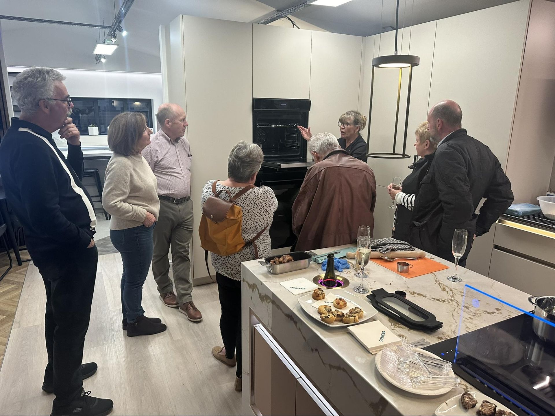 Group of people in a modern kitchen, viewing an oven. Someone points to the appliance, with snacks on the island.