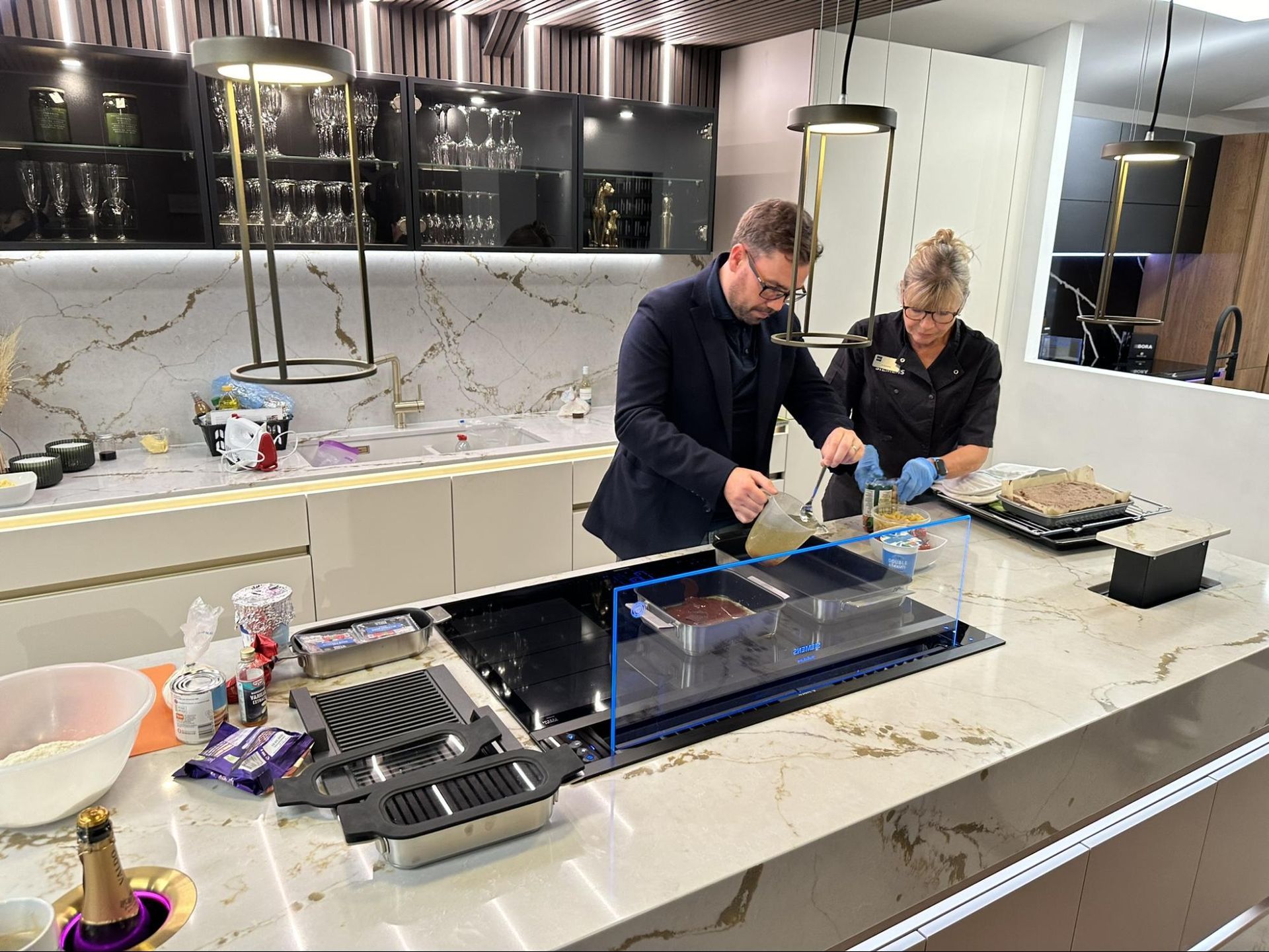 Two people cooking at a kitchen island. One pours liquid into a pan set on a cooktop. Luxurious kitchen.