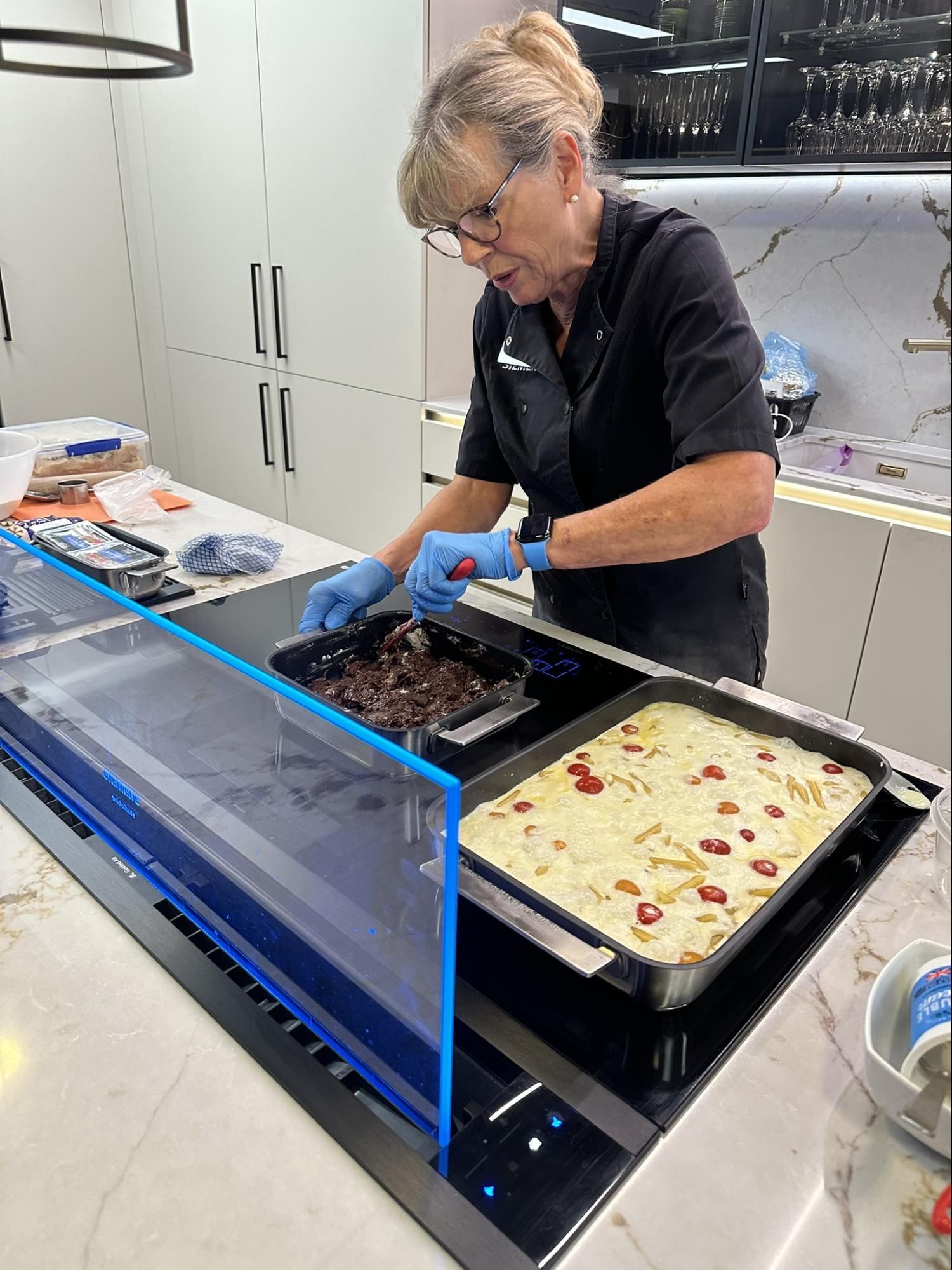 Woman cooking in kitchen, using spatula. Two trays on stove, one with dark food, one with white. Blue gloves.