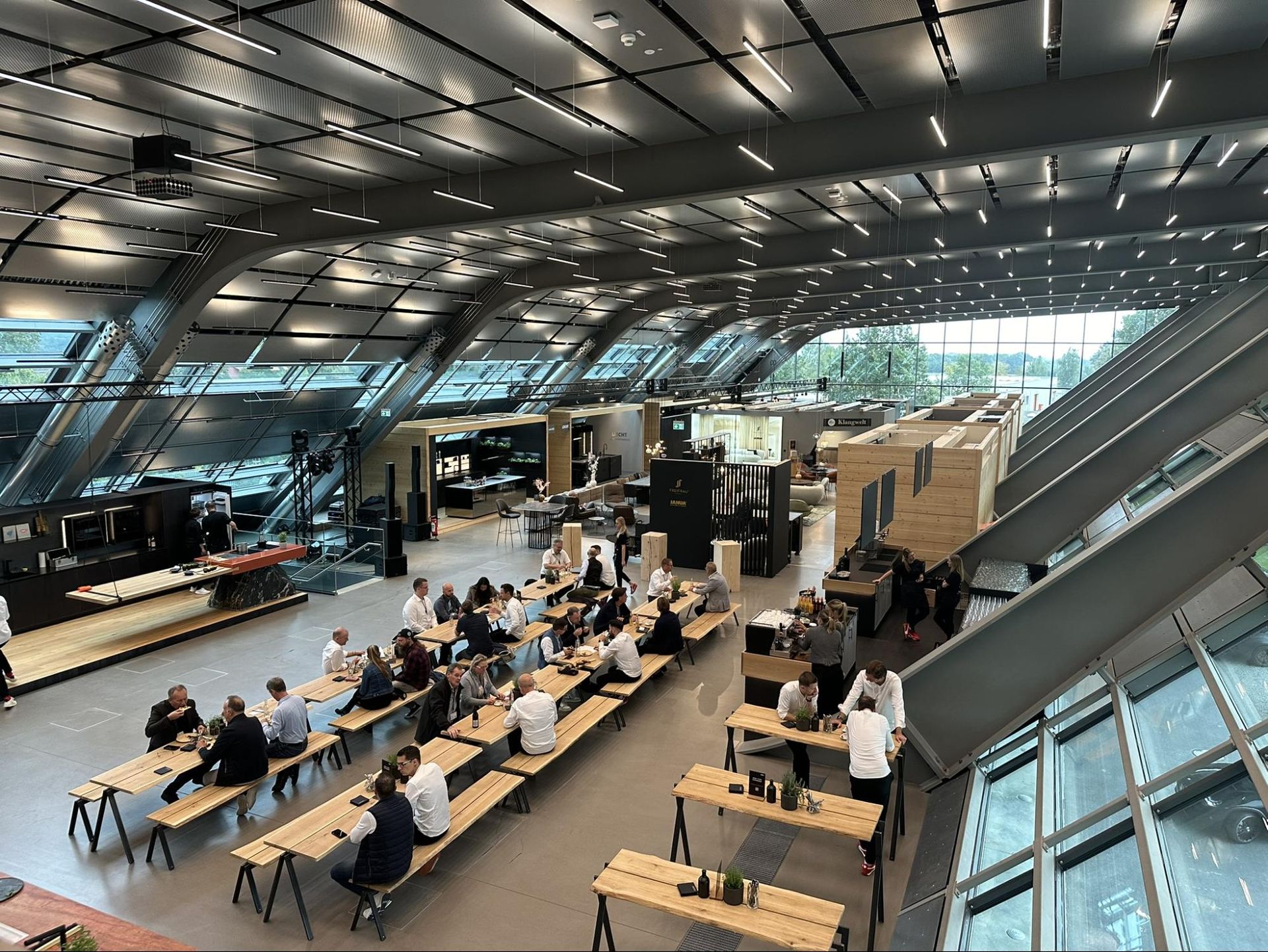 Large event space, people seated at long tables, dark furnishings, glass roof, modern architecture.