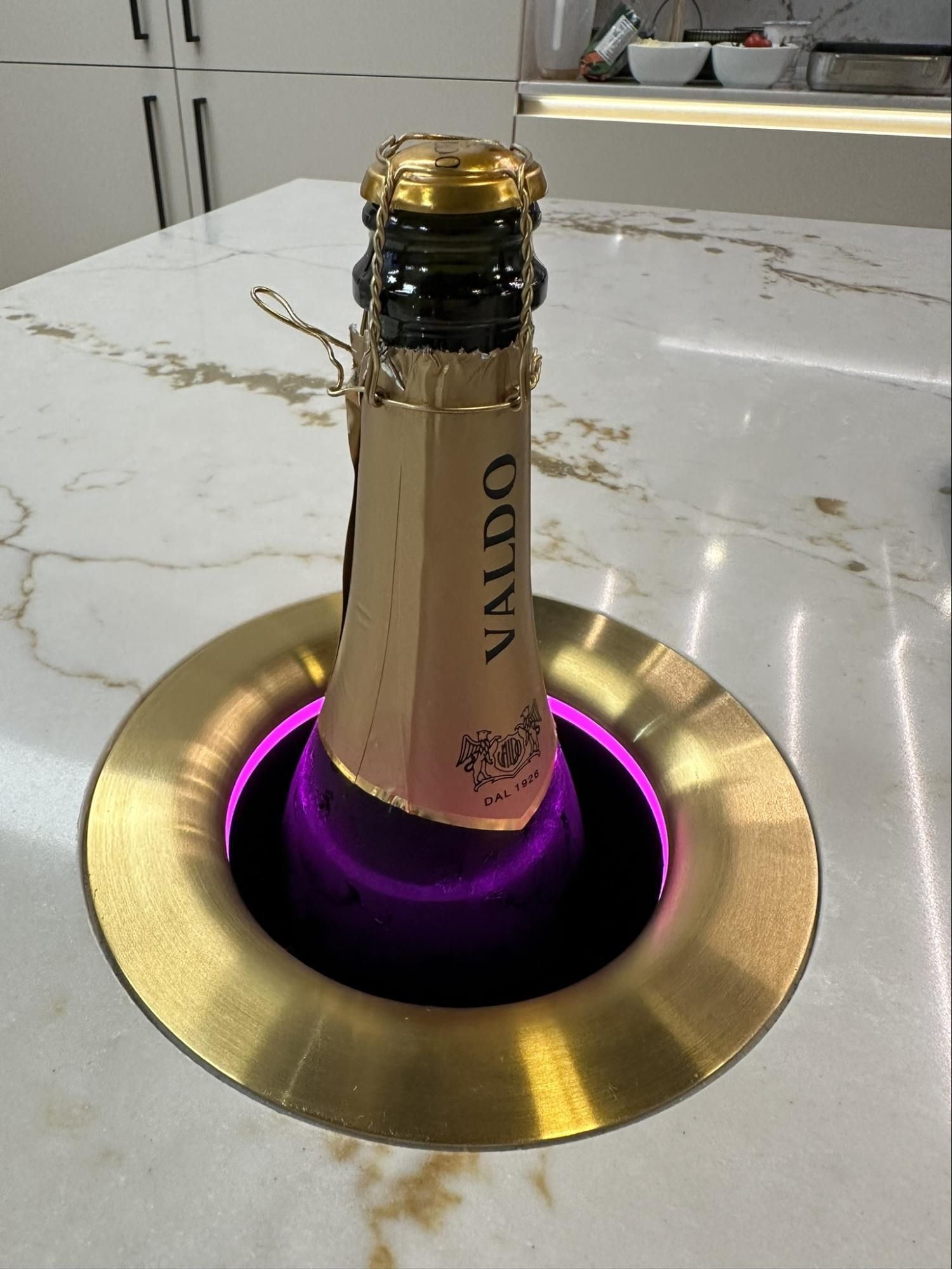 Champagne bottle in a brass-rimmed, lit cooler on a marble countertop. The light inside glows purple.