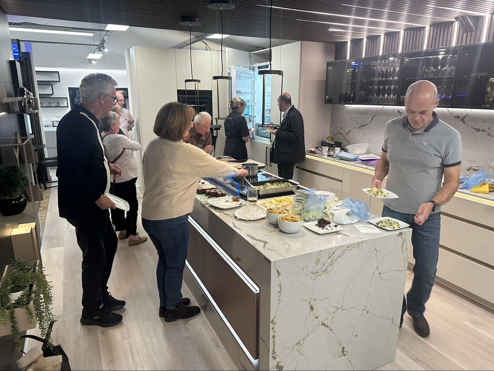 People at a kitchen showroom buffet, selecting food from a countertop island.