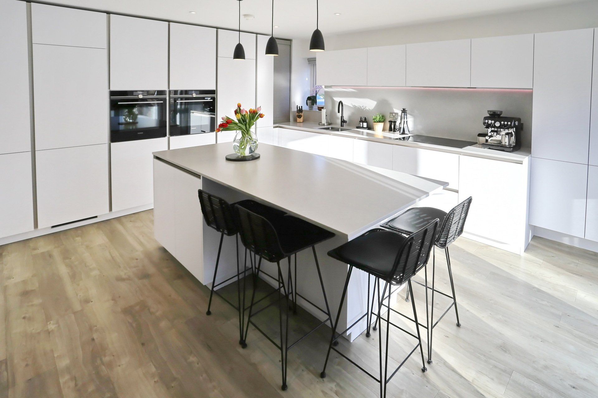 Modern white kitchen with island, black stools, and overhead lighting. Wooden floors and large windows.