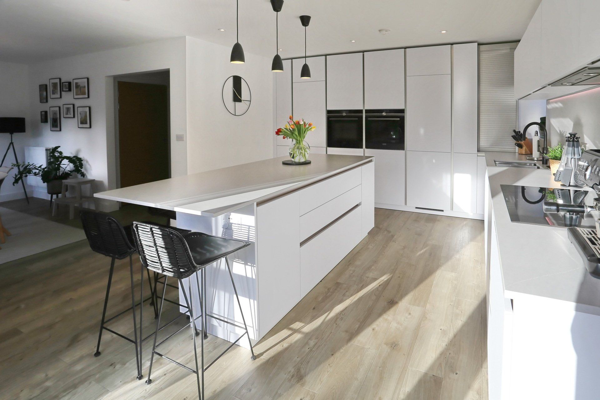 Modern white kitchen with island, bar stools, and overhead lighting. Sunlight streams in.