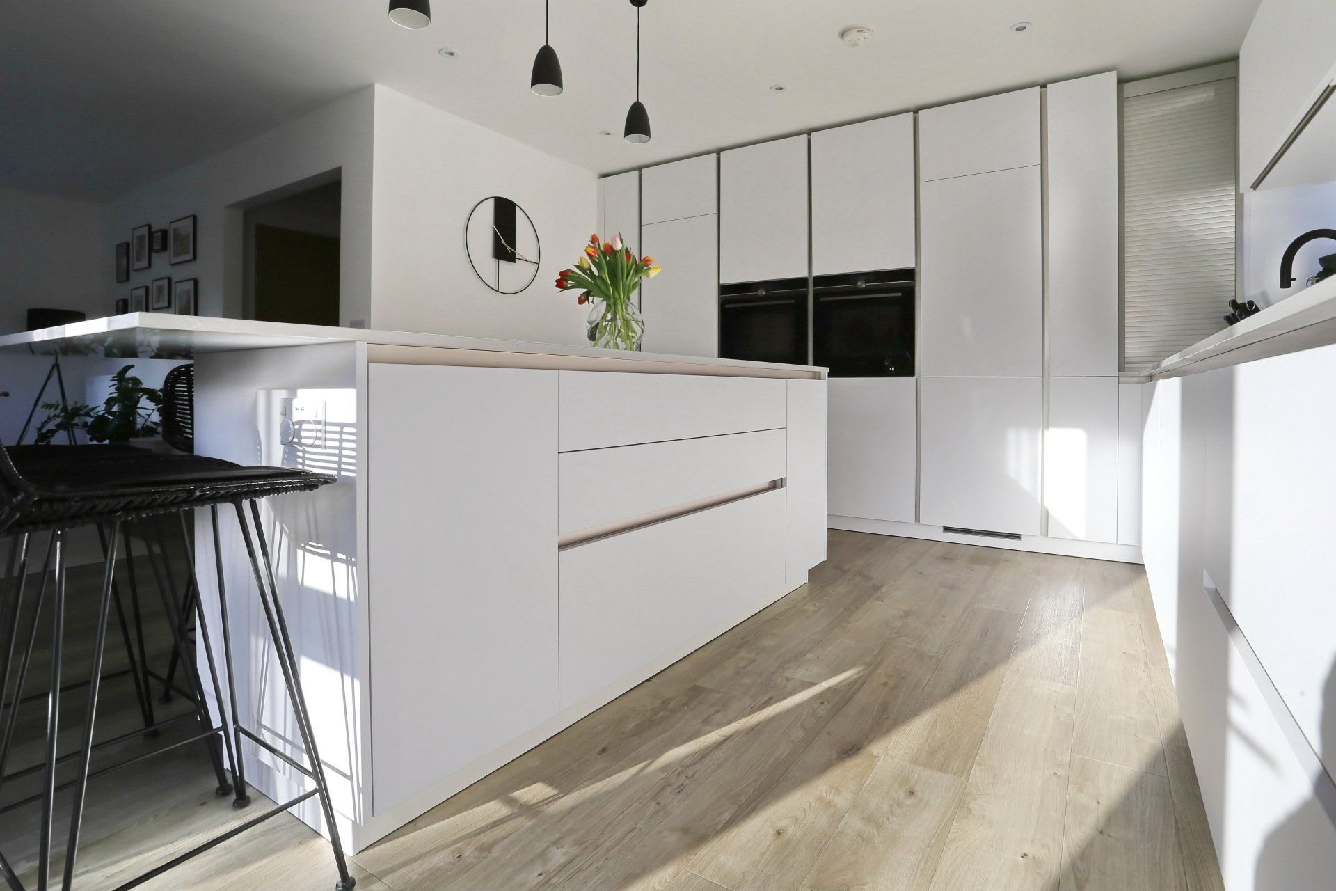 Modern white kitchen with island, wooden floors, black stools, and black oven.