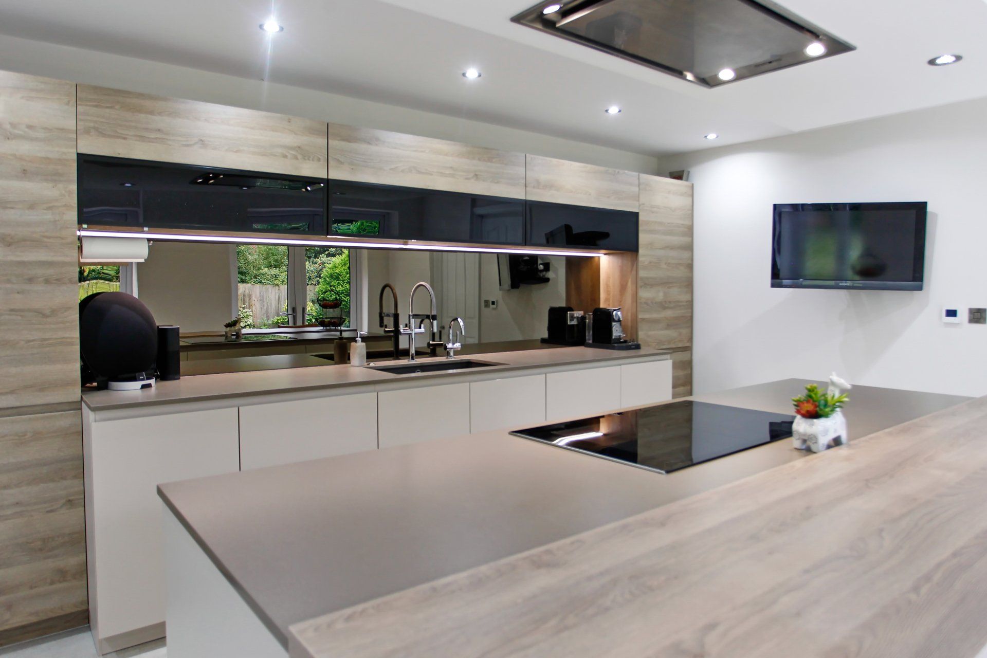 Modern kitchen with light wood cabinetry, grey countertops, island with stovetop, black accents, and a mounted TV.