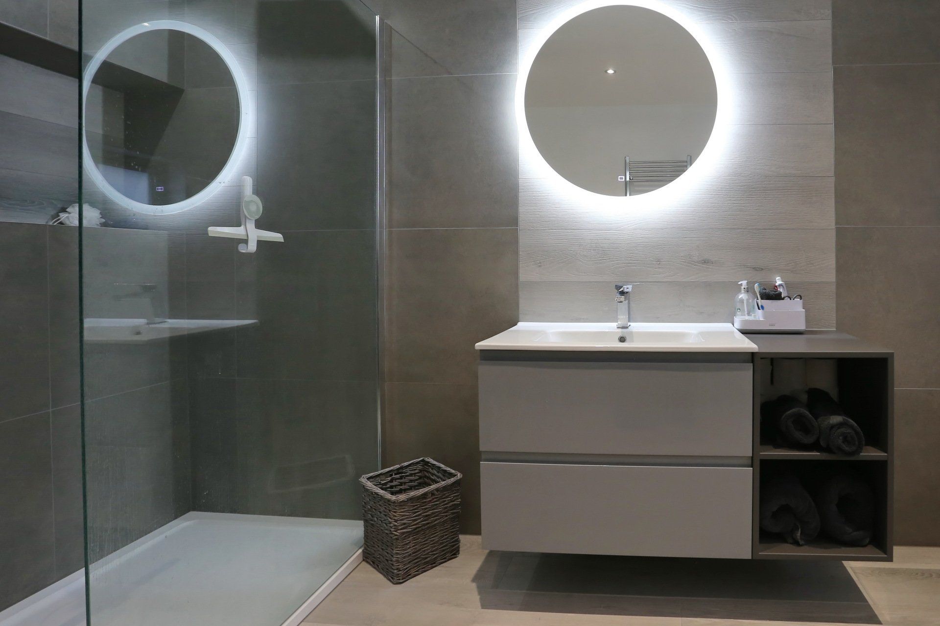 Modern gray bathroom with shower, floating vanity, and round mirrors with backlights.