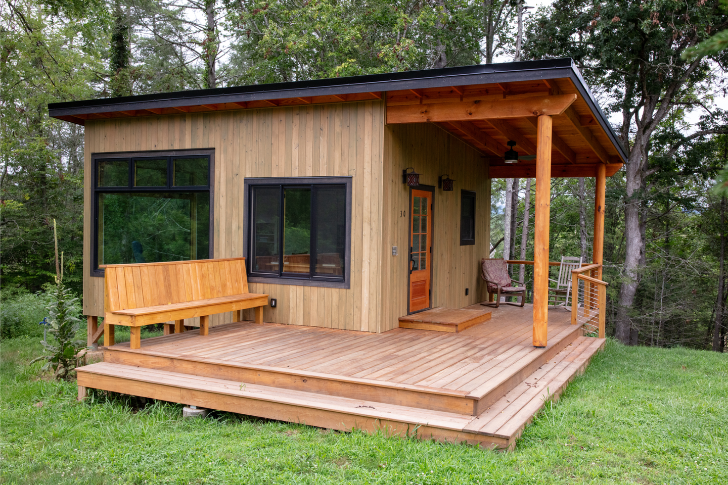 A modern wooden house with a dark roof and wraparound deck in a lush green setting.