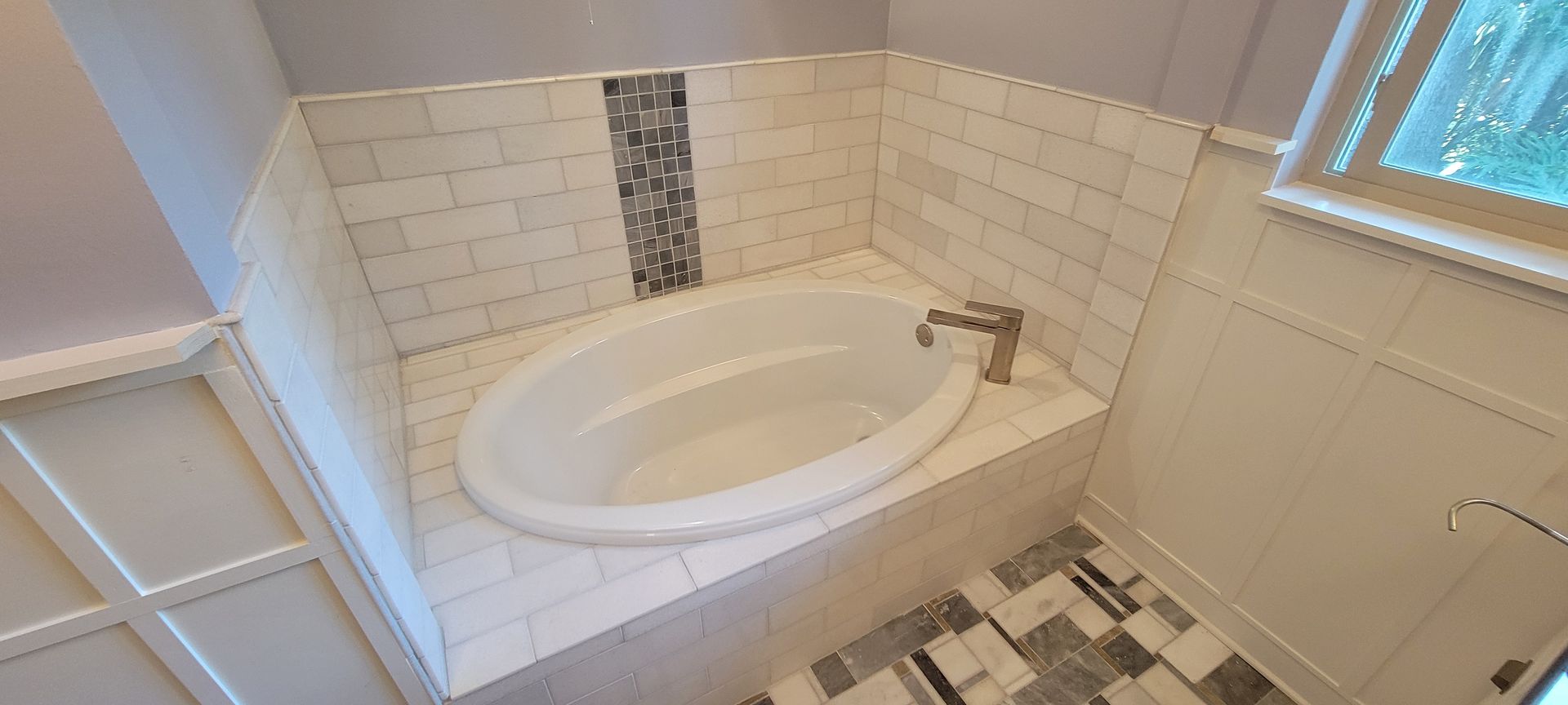 An oval white bathtub set in a tiled alcove with white subway tile walls, a decorative mosaic accent, and marble floors.