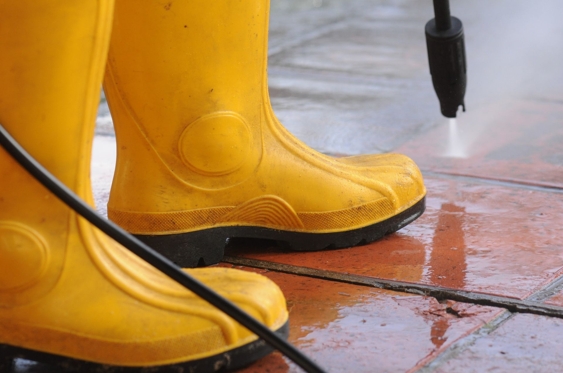 A person in bright yellow rubber boots uses a pressure washer to clean a wet, tiled patio.