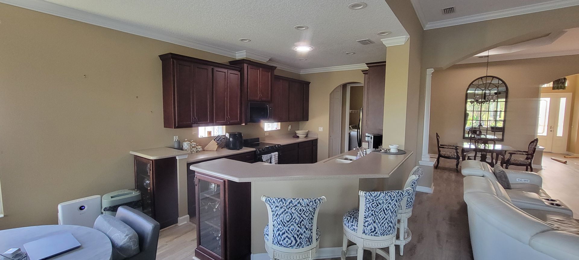 A spacious kitchen with dark wood cabinets, a tan island with two patterned chairs, and a view into a dining room.