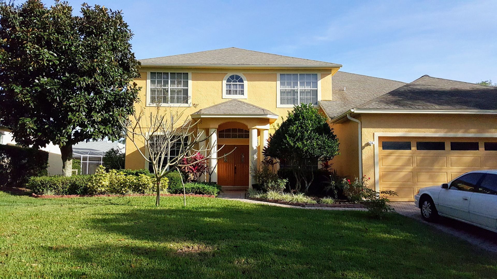 A two-story yellow house with a gabled entrance, a single garage, and a large tree in the front yard on a sunny day.