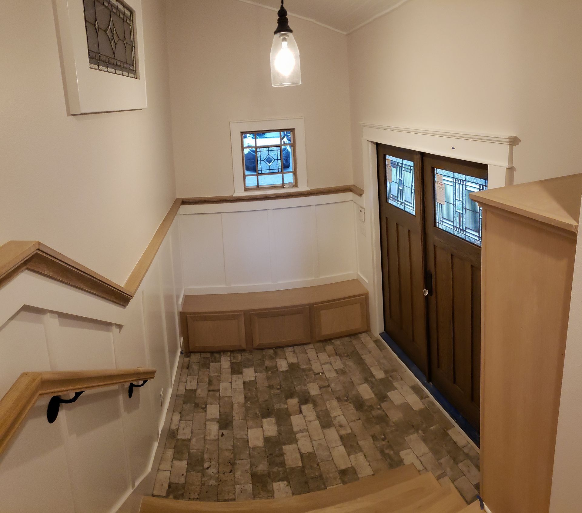 A bright entryway with brick flooring, a built-in wooden bench, a pendant light, and a double wooden door with glass.