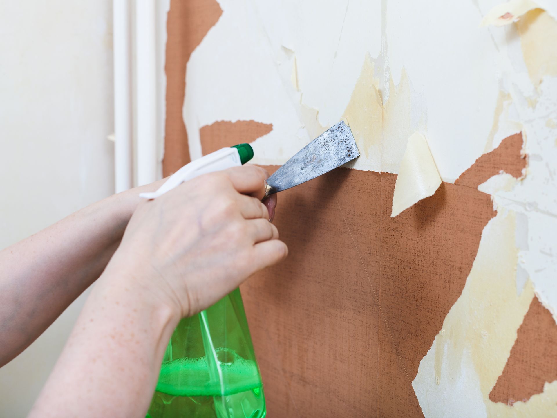 A person is removing wallpaper from a wall with a spatula and spray bottle.