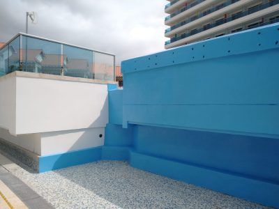Blue rooftop terrace beside a white wall and apartment building under cloudy skies