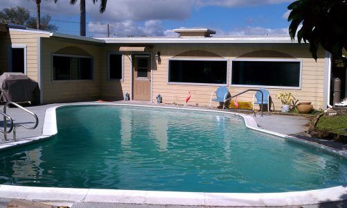 Backyard swimming pool with a house and patio chairs under a partly cloudy sky