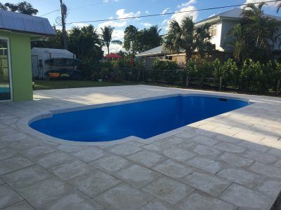 Backyard swimming pool with blue water and a stone deck, surrounded by tropical plants and houses