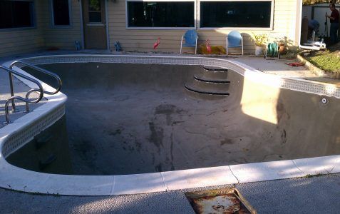 Empty abandoned swimming pool in a backyard, with faded concrete and a diving platform.