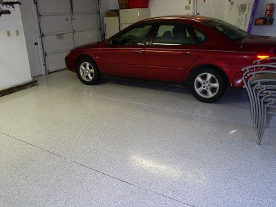 Red car parked in a garage with a speckled white floor and stacked chairs on the right.