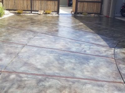 Sunlit concrete driveway with dark stain patterns leading to a wooden gate and garage area