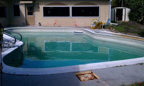 Backyard swimming pool with turquoise water beside a house and patio, cover partially rolled up on the left