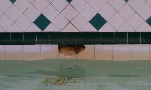 Indoor pool wall with white tiles, green diamond accents, and a damaged section near the waterline.