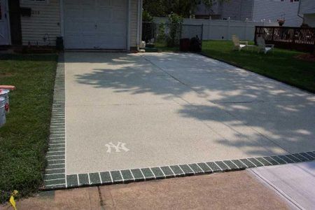 Concrete driveway with green brick border beside a suburban house, partly shaded by trees.
