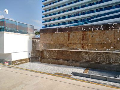 Concrete wall beside a sidewalk, with a modern blue-and-white building in the background