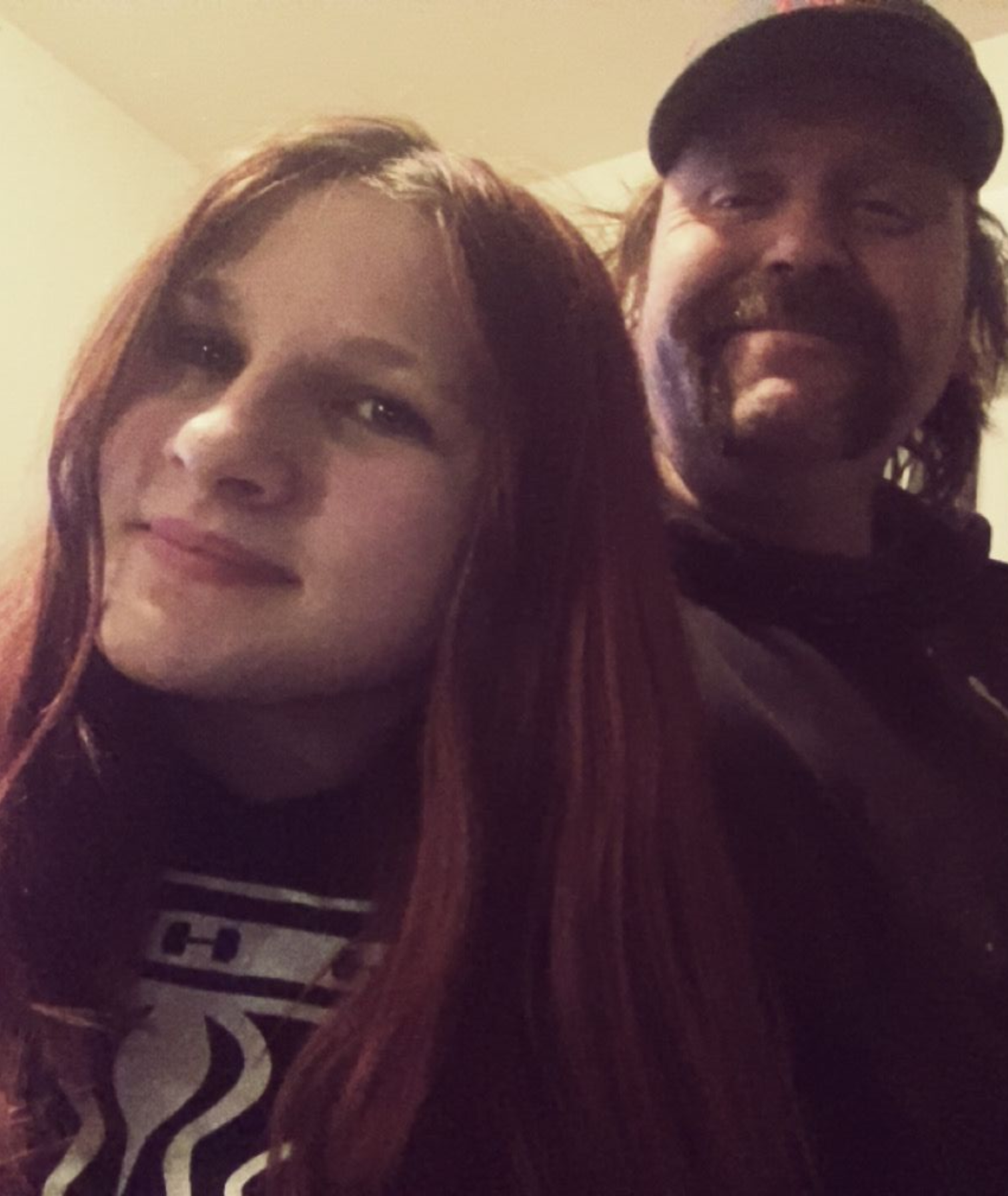 Two people posing closely together indoors, one in front with long brown hair and a slight smile.