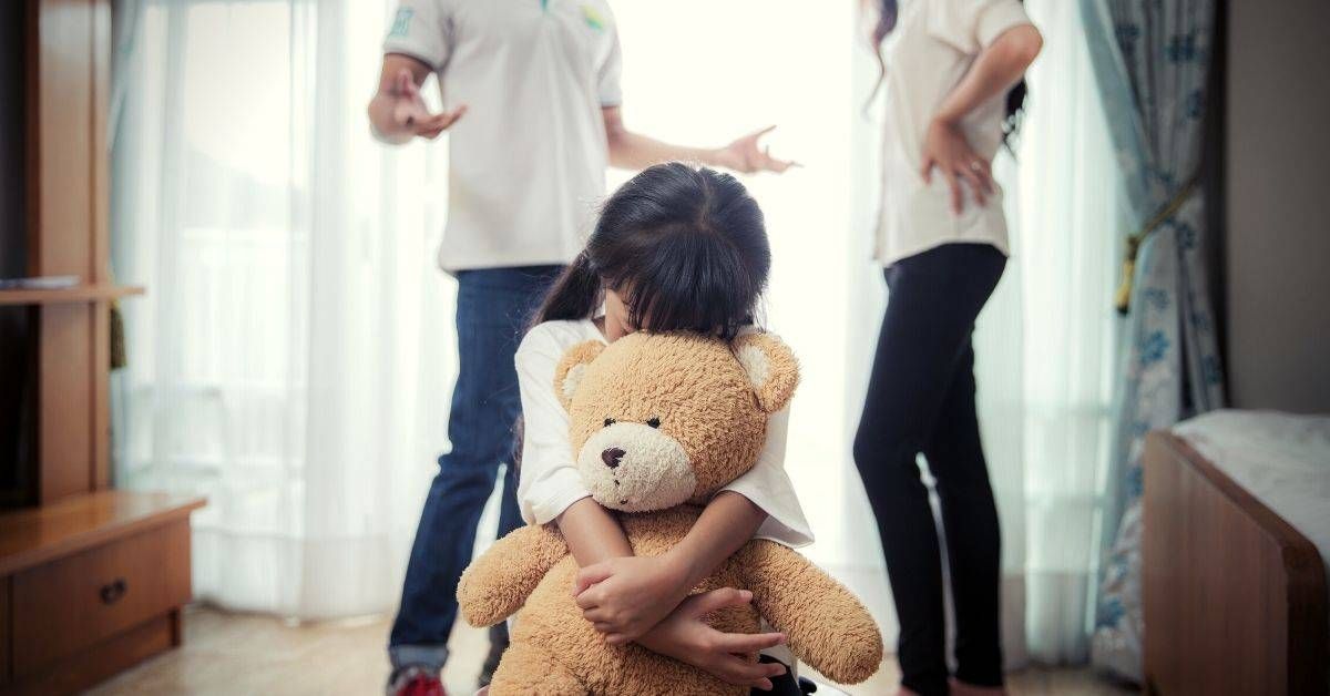 A child is holding a teddy bear while a man and woman argue in the background.
