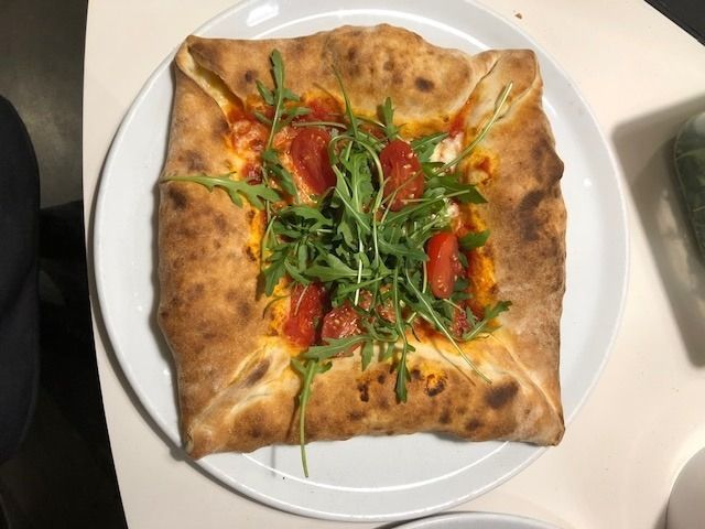 Pizza quadrata con base ripiegata, condita con pomodori e rucola su un piatto bianco.