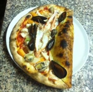 Mezza pizza ai frutti di mare su un piatto bianco; la crosta è scura e frizzante, guarnita con cozze, granchio e verdure.