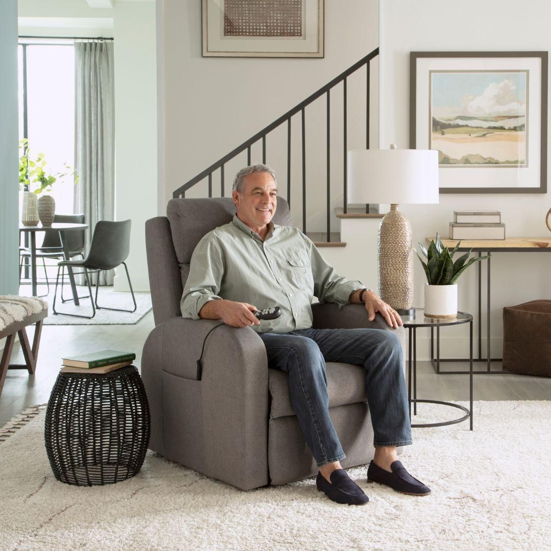 Man seated in a gray lift recliner, holding remote. Living room setting with table, lamp, and art.