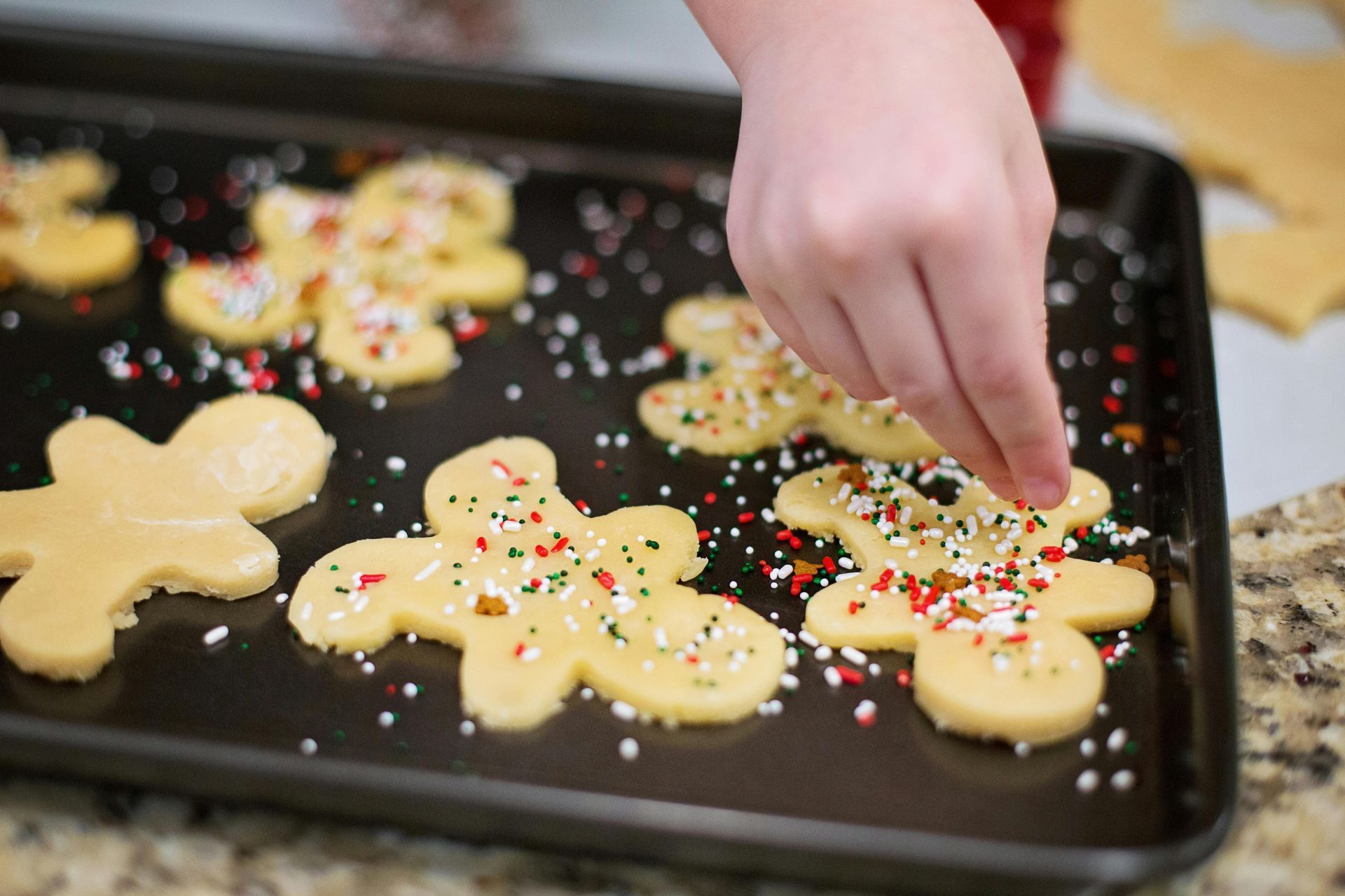 Child decorating gingerbread cookies with colorful sprinkles on a baking sheet.