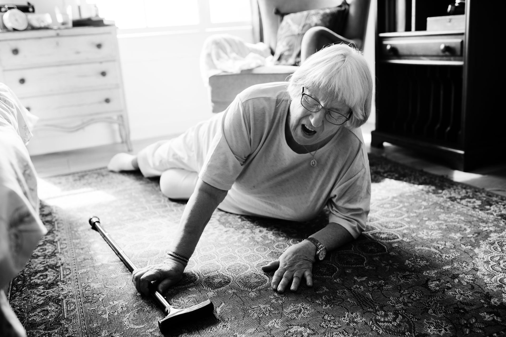 Elderly woman on the floor, cane nearby. She is wearing glasses, appears distressed, in a bedroom.