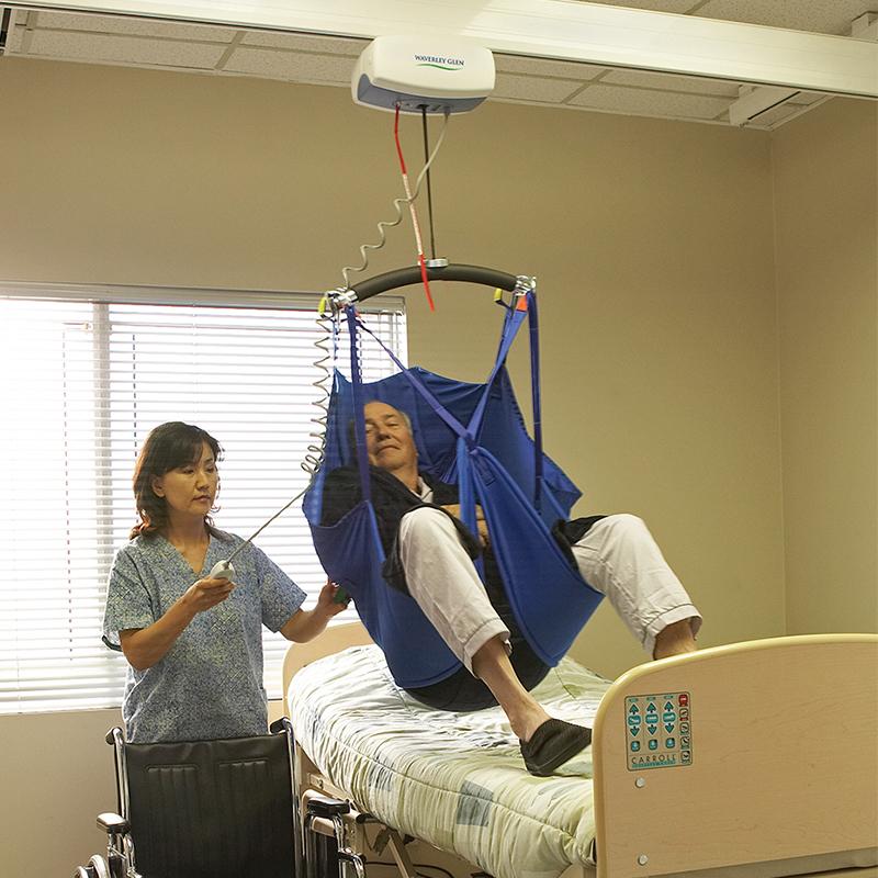 Woman assisting a person in a sling lift. The person is sitting in a sling above a bed.