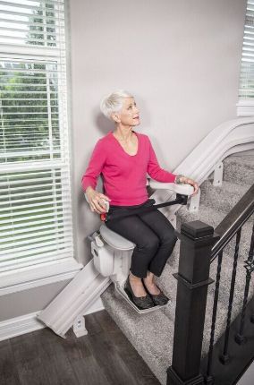 Woman in a stairlift ascends stairs. She's wearing a pink shirt and black pants. The lift is white, the stairs are carpeted.
