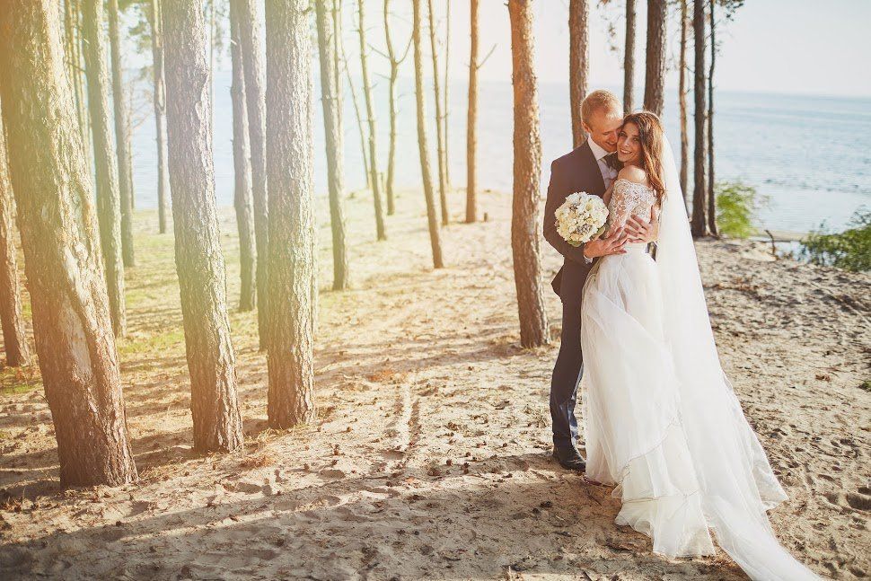 Couple embraces, bride in white gown, groom in suit, standing in a sunlit forest near water.
