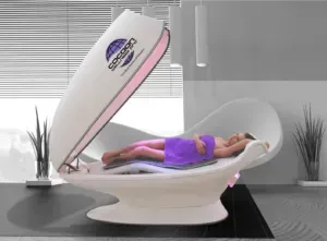 A person reclines inside a white Cocoon Wellness Pod, lid open. Purple towel, grayscale room.