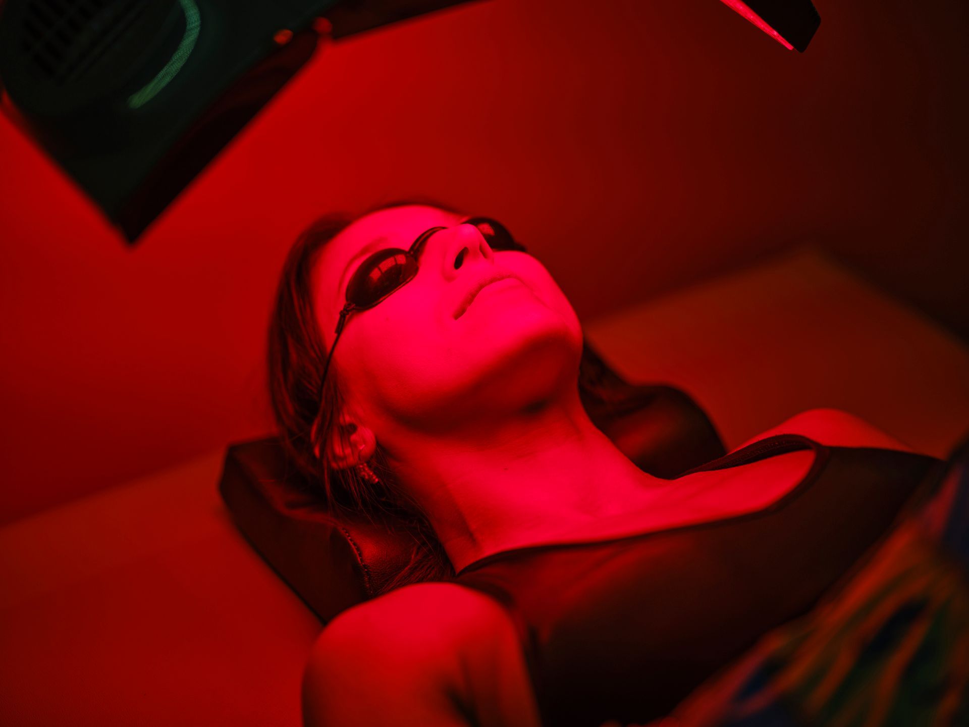 Woman wearing eye protection lies under red light therapy, lit in red. Woman wearing eye protection lies under red light therapy, lit in red.