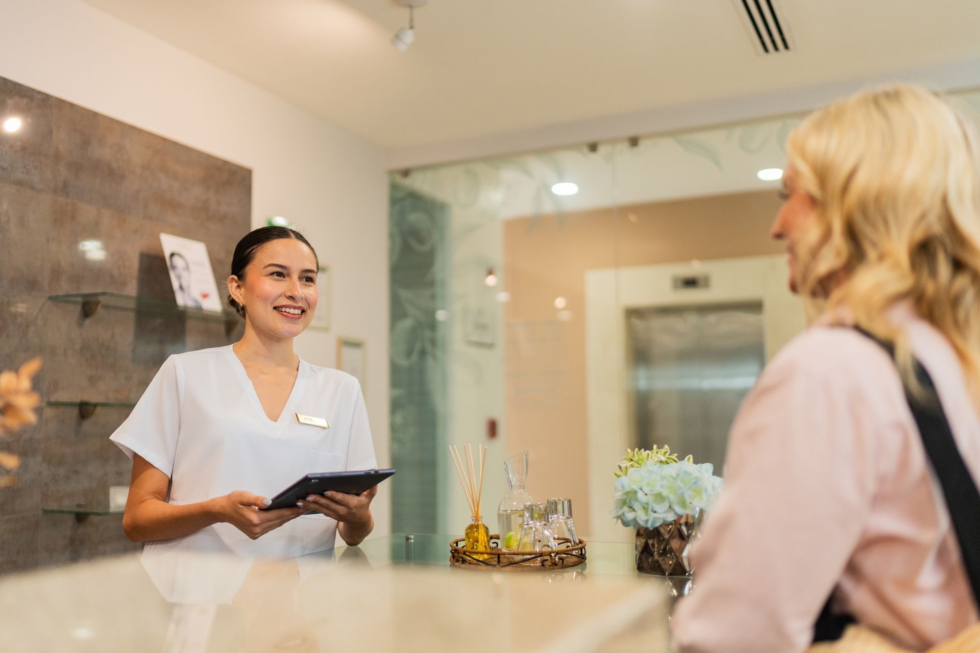 Receptionist in white uniform smiling, talking to a customer at a spa reception desk.