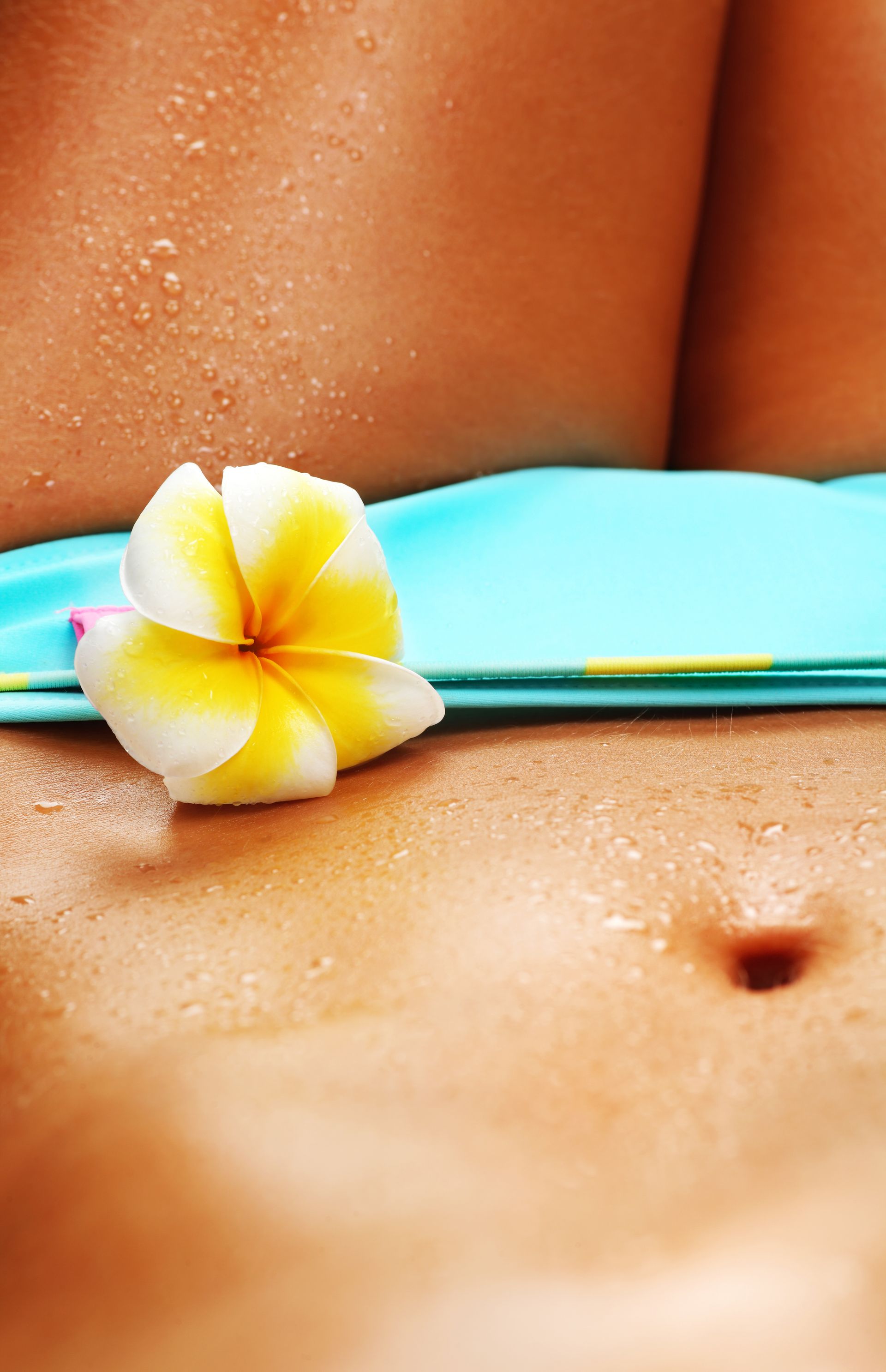 Close-up of a person's sun-kissed torso, a blue bikini top, and a white and yellow frangipani flower.