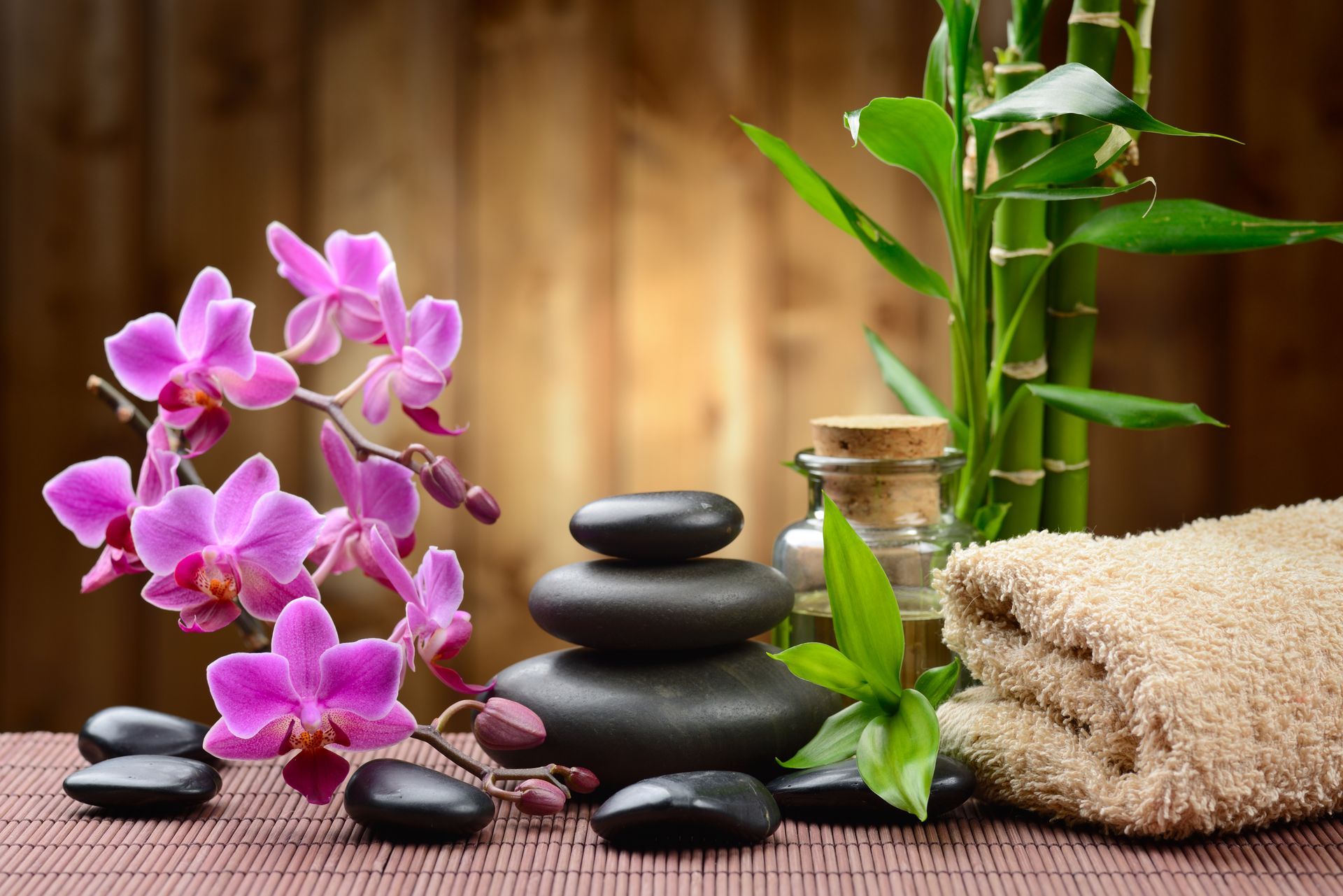Stack of smooth black stones, purple orchids, bamboo, towel, and oil bottle arranged on bamboo mat.