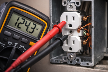 A multimeter probe tests a wall outlet, displaying 124.3 volts on its screen.