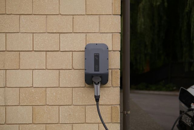 A gray electric vehicle charging station mounted on a beige brick wall with a charging cable plugged into its base.