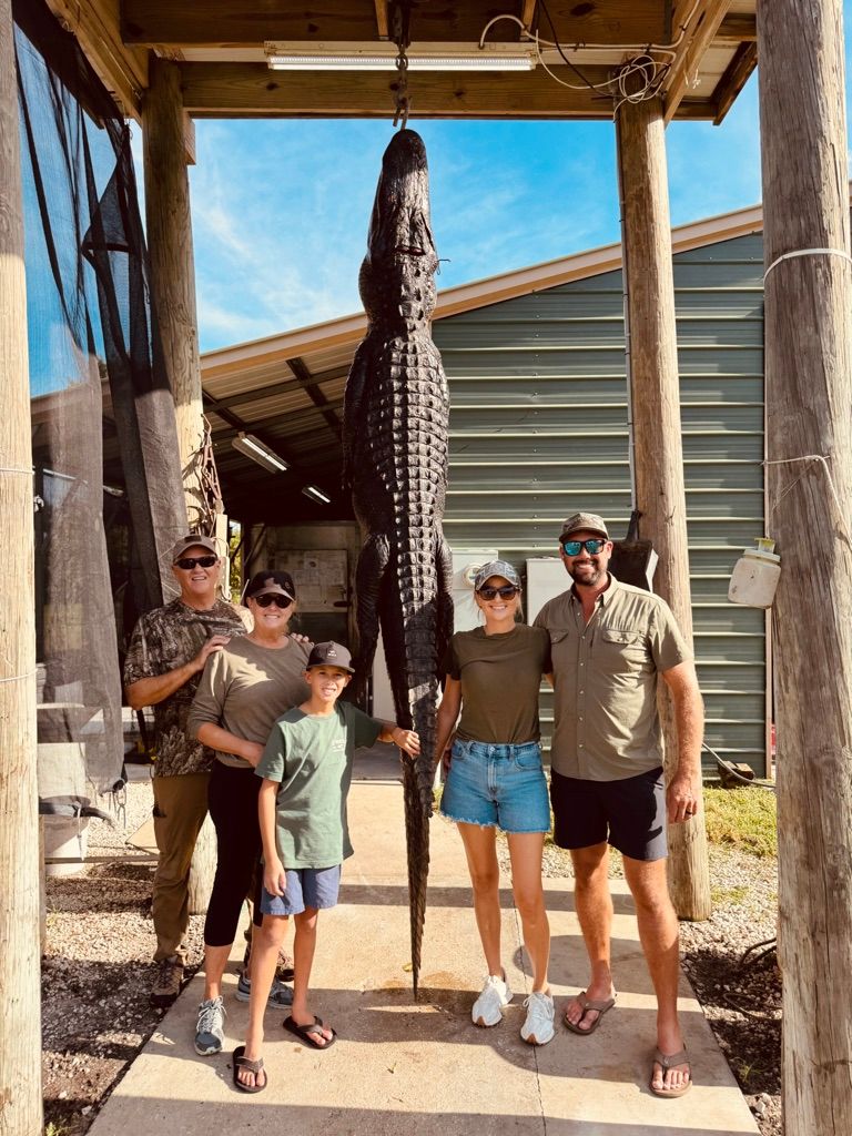 Group of five people posing beneath a large alligator hung from a processing shed hoist