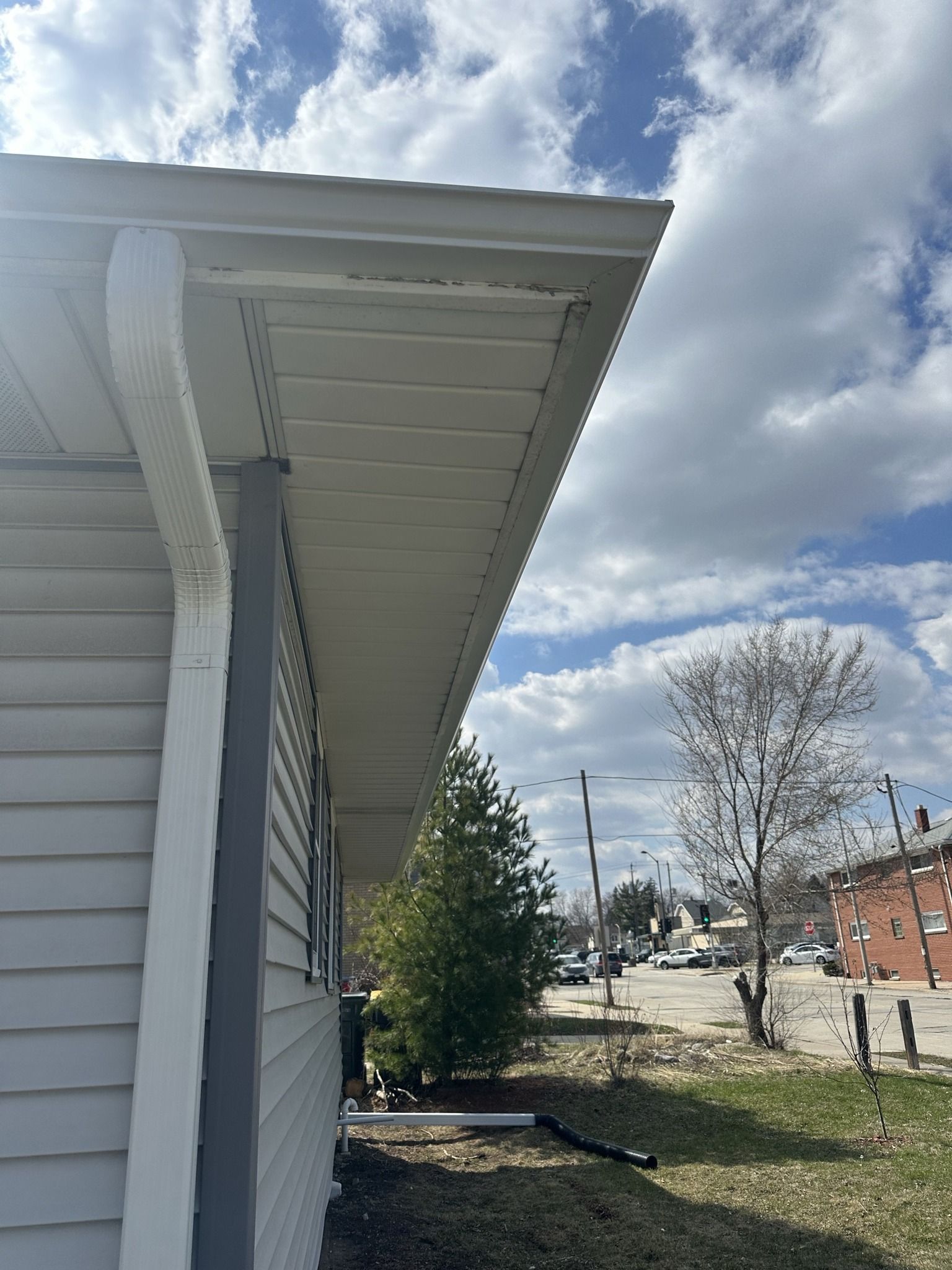 Exterior view of the corner of a house with grey siding, a white downspout, and a soffit under a roof overhang.